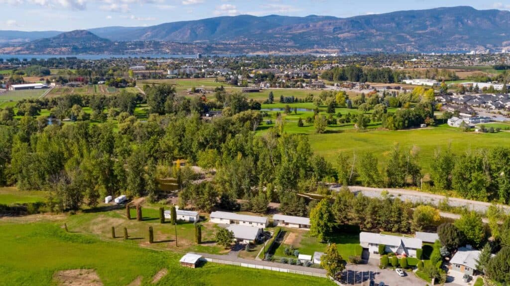 Aerial view of a lush green property in Kelowna, BC, showcasing farmland, residential homes, and scenic mountain views in the background.